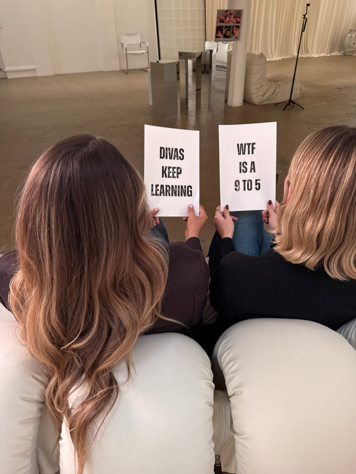 Annie and Kate seated with their backs to the camera, holding cards reading “Divas keep learning” and “WTF is a 9 to 5”