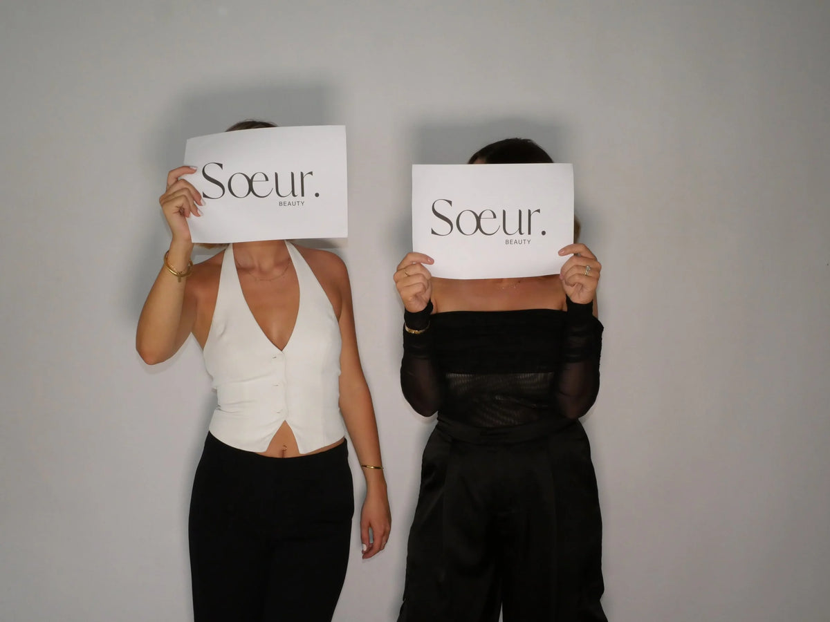 Annie and Kate standing side by side against a pale wall, each holding a Sœur. Beauty card in front of her face
