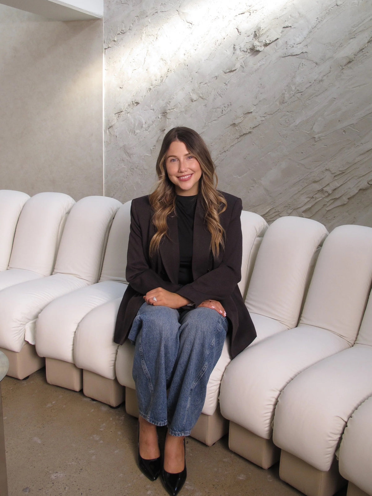Annie Kelly-Boxall, owner of Sœur. Beauty, seated on a white curved sofa in front of a textured stone wall